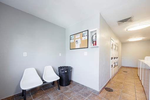 Two white chairs are placed in a room with a trash can and a bulletin board.