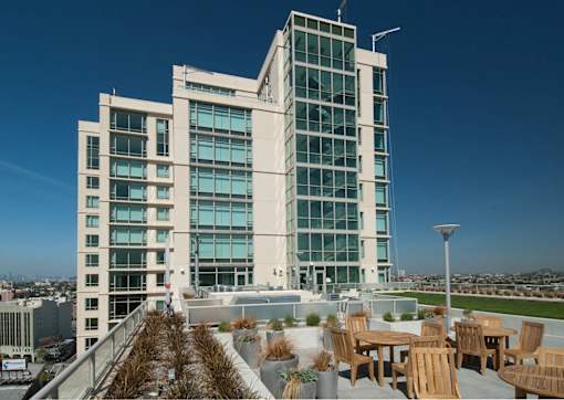A modern building with a glass facade and a rooftop patio with wooden furniture at The Grand Apartments, California