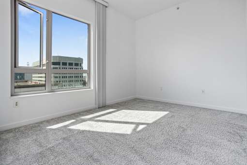 A large empty room with a carpeted floor and a large window at The Grand Apartments, Oakland, CA