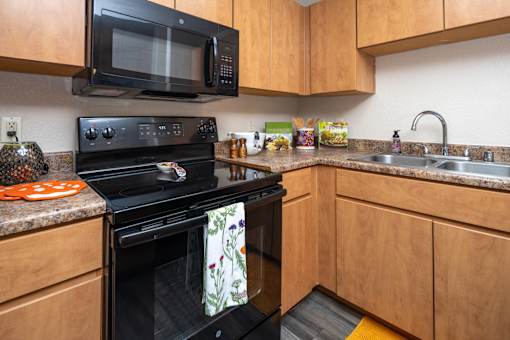 Surprise, AZ, Apartments for Rent - The Village at West Point - A Kitchen With Wood Cabinets, Black Stove And Microwave, Double Sink, And Speckled Countertops.