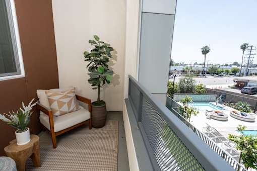 A chair with a pillow and a potted plant sit on a balcony overlooking a pool.