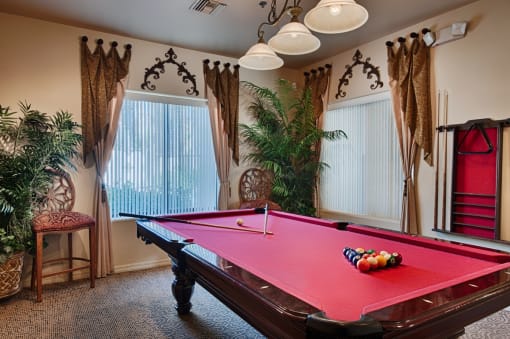 Surprise, AZ Apartments for Rent - The Village at West Point - Billiards Room with Carpeted Flooring, Window Drapes, Houseplants, Chairs, Hanging Light Fixtures, Billiards Table, and Windows with Vertical Blinds.