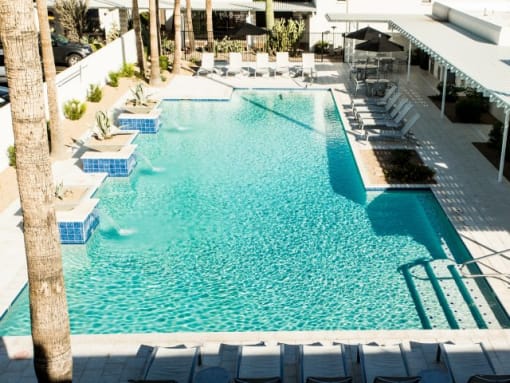 Apartments in Scottsdale, AZ - Resort-Style Swimming Pool, With Lounge Chairs, Tables With Umbrellas, Well-Kept Landscaping, Water Fountains, And Palm Trees.