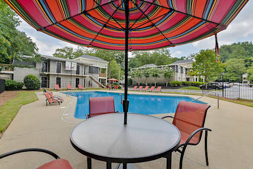 A table with chairs and an umbrella is in the foreground of a pool area.