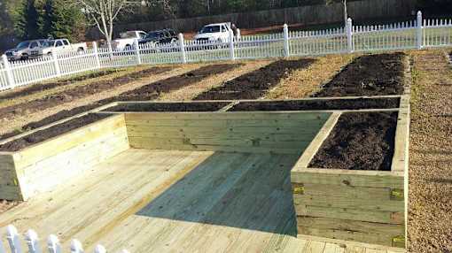 A wooden raised garden bed with soil in the foreground and a white picket fence in the background.