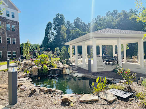 A gazebo is situated in a garden with a pond and a building in the background.