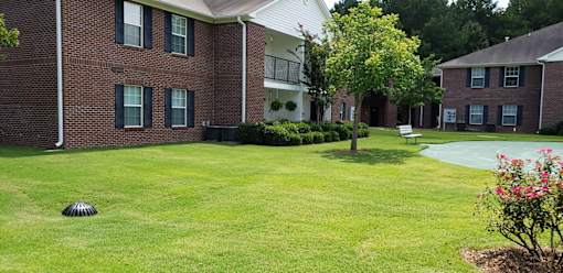 A grassy area in front of a brick building with a tree and a flower bed.