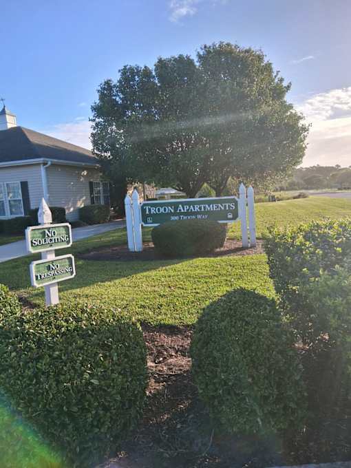 A sign for Troon Apartments stands in front of a house.