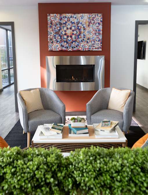 A living room with two grey chairs, a coffee table with books and a fireplace with a painting above it.
