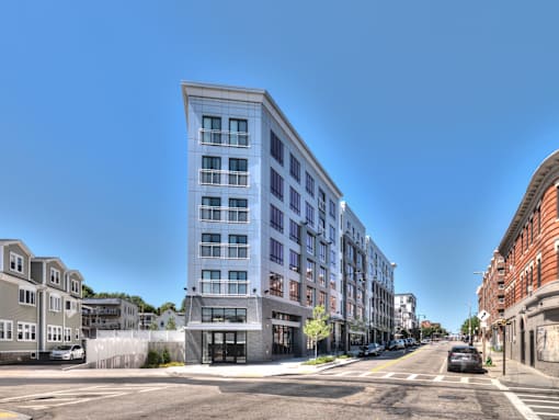 A street view of a city with a large building on the left and a car driving on the road.