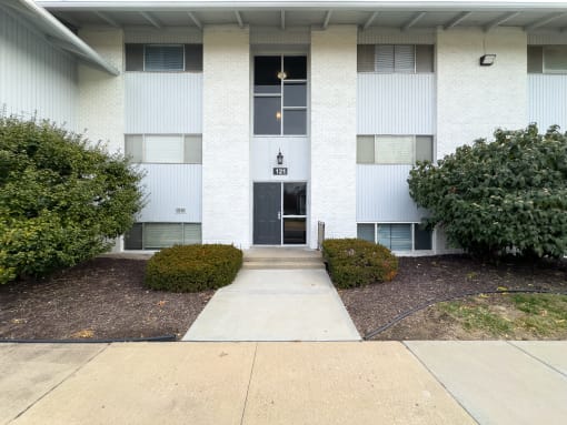 the front entrance to a white building with bushes and a sidewalk