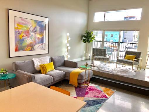 A living room with a grey couch, yellow pillows, a yellow blanket, a small table with a plant, a chair with a yellow cushion, and a painting on the wall.