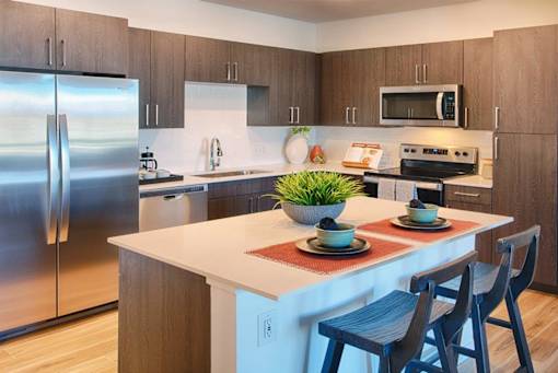 A modern kitchen with a stainless steel refrigerator, wooden cabinets, and a white island with a potted plant on top.