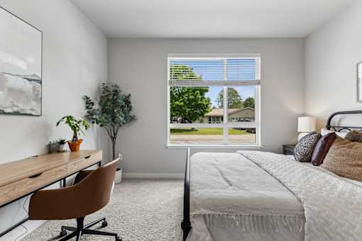 A bedroom with a bed, desk, chair, and a window overlooking a green outdoor area.