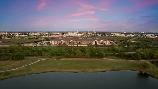 A large building complex is surrounded by a grassy field and a body of water under a pink and blue sky.