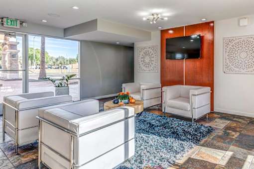 A living room with a white couch, a white chair, a white ottoman, a blue rug, a wooden cabinet, a flat screen TV, and a window with a view of the outside.