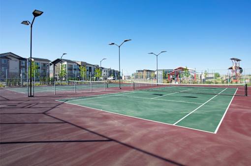 A tennis court with a red surface and white lines, surrounded by a fence and buildings in the background.