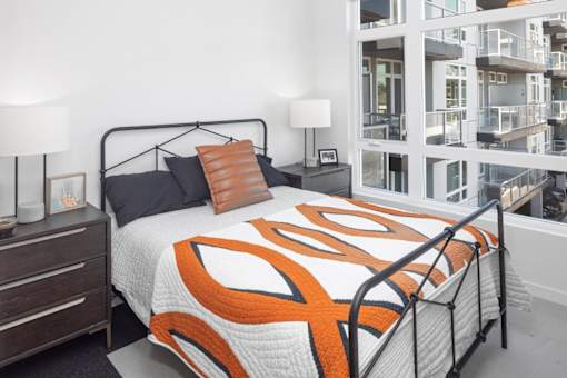A bed with a black frame and an orange and white comforter is in the foreground of a room with a white wall and a window.