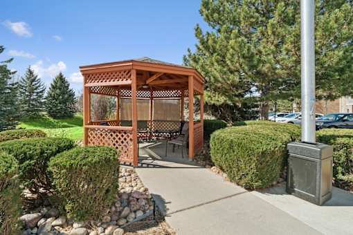 A wooden gazebo is surrounded by green bushes and a stone wall.