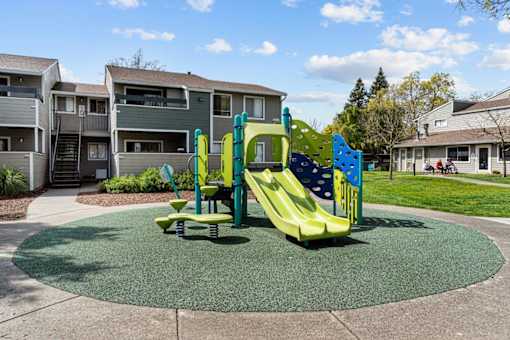 A playground with a green and yellow slide in the middle of a grassy area.