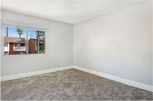 A room with a carpeted floor and a window showing a view of a house and trees outside.
