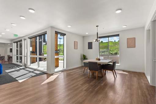 A room with a dining table and chairs in the foreground and a view of the outside through the windows.