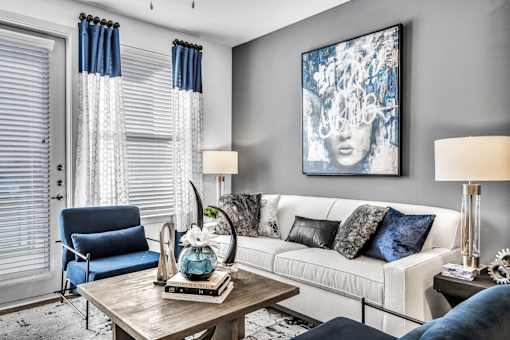 A living room with a white couch, blue chair, and a painting on the wall.