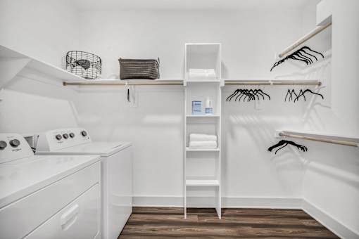 A white laundry room with a washer and dryer, a shelf with clothes, and a rack with clothes hangers.