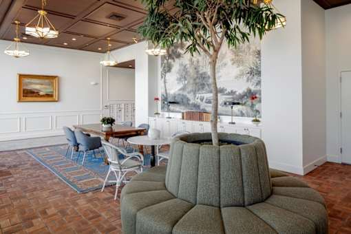 A tree in a pot sits on a round ottoman in a room with a painting on the wall.