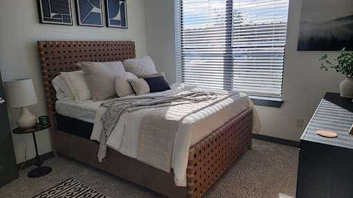 A bedroom with a bed, nightstand, and window with blinds at Urban Square, Denton, Texas