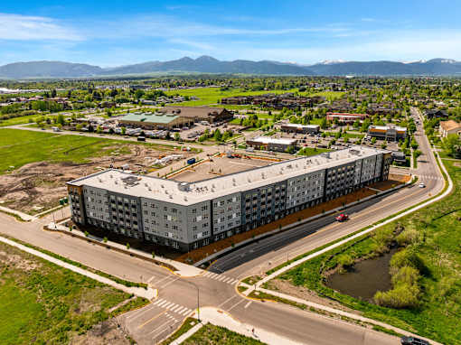 A large grey building is surrounded by a grassy area and a road with cars at The Beaumont Apartments, Bozeman, MT