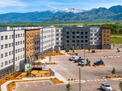 A parking lot in front of apartment buildings with a playground and mountains in the background at The Beaumont Apartments, MT 59715
