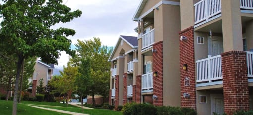 View of Country Oaks apartment building 4 with red brick, white beam balconies, and manicured grass pathways with trees at Country Oaks Apartments in Clearfield, Utah 84015