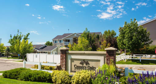 Country Oaks welcome area with manicured landscaping and branded signage in front of apartment homes with a blue sky and small white clouds at Country Oaks Apartments in Clearfield, Utah 84015