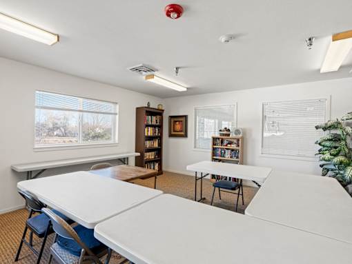 A room with a white table and chairs, a bookshelf, and a window with a view of trees.