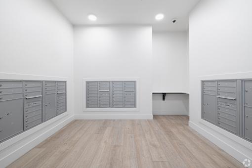 A room with white walls and wooden flooring, featuring a row of grey lockers on the left and a bench on the right.