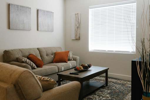 A living room with a grey couch, a brown armchair, a coffee table with a bowl of fruit on it, and two abstract paintings on the wall.