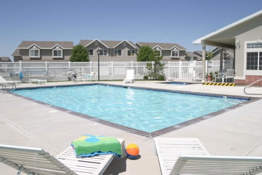View of the blue swimming pool with a white fence, white chaise lounge chairs with a colorful towel on one and a multicolored beach ball in between, and resident building to the right at Oakstone Apar