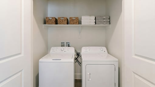 A white laundry room with two washing machines and a shelf with baskets and towels.