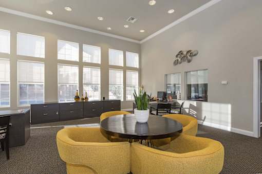 A round table with a plant on it is surrounded by yellow chairs in a room with large windows.