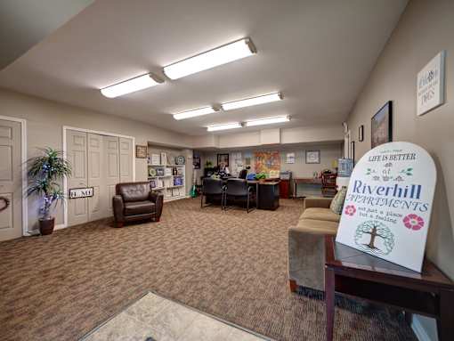 A sign in the foreground of a room with a carpeted floor and a few chairs.