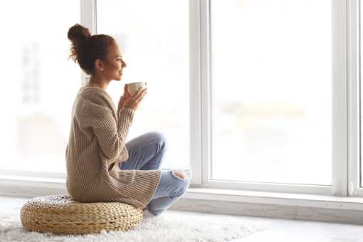 A woman in a beige sweater is sitting on a woven stool by a window.