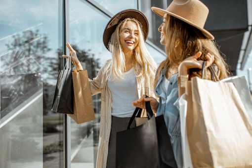 Two women wearing hats and carrying shopping bags are standing outside a store.