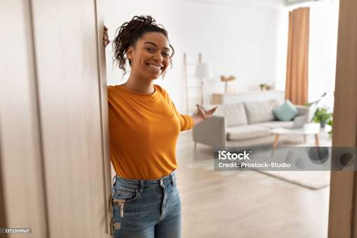 A woman in a yellow top and blue jeans stands in a room with a white wall and a couch in the background.