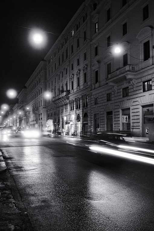 A black and white image of a city street at night with cars passing by.