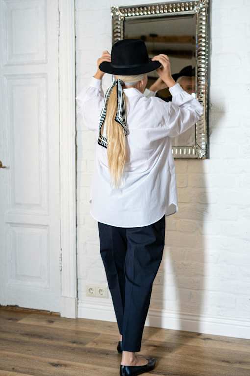 A person in a white shirt and black pants is adjusting a black hat in front of a mirror.