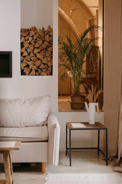 A living room with a white couch, a small table with a plant on it, and a picture of wood stacked on the wall.