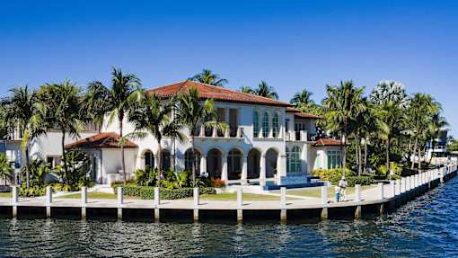 A large white house with a red roof is situated on a dock with palm trees in the background.