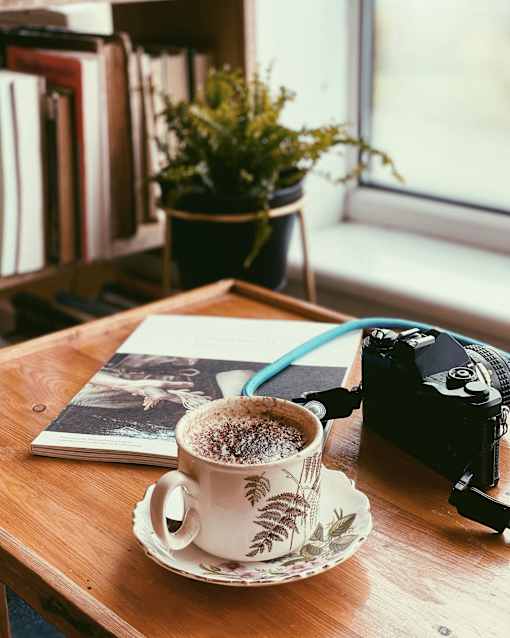 A cup of coffee sits on a saucer next to a camera and a book on a wooden table.