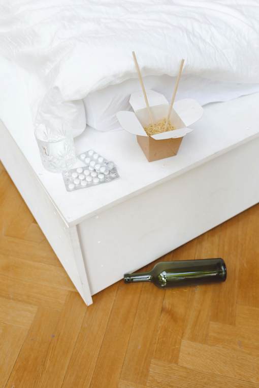 A bottle of wine is on a wooden floor next to a bed.